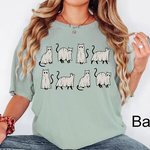 May include: A light green t-shirt with a graphic of nine black cats dressed as ghosts. The text "Bay" is in the bottom right corner of the image.