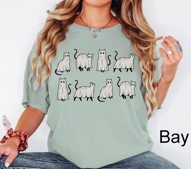 May include: A light green t-shirt with a graphic of nine black cats dressed as ghosts. The text "Bay" is in the bottom right corner of the image.