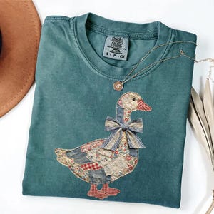 Peut inclure: T-shirt Comfort Colors bleu sarcelle avec un motif d'oie en patchwork. L'oie a un n&oelig;ud bleu et est faite de divers tissus &agrave; motifs. Un collier en or avec un petit pendentif est drap&eacute; sur le t-shirt.