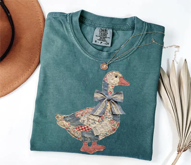 Peut inclure: T-shirt Comfort Colors bleu sarcelle avec un motif d'oie en patchwork. L'oie a un n&oelig;ud bleu et est faite de divers tissus &agrave; motifs. Un collier en or avec un petit pendentif est drap&eacute; sur le t-shirt.