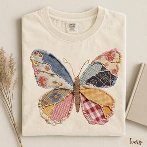 May include: Ivory-colored t-shirt featuring a large, colorful butterfly applique. The butterfly wings are made of various patterned fabrics, including floral, denim, and plaid. The shirt has a crew neck and short sleeves. The tag reads "Comfort Colors".