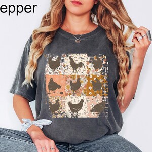 May include: Dark gray t-shirt with a patchwork design featuring chicken silhouettes on a floral background. The word "Pepper" is printed in the upper left corner. The shirt has short sleeves and a relaxed fit.
