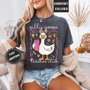 Puede incluir: Camiseta gris oscuro con un gr&aacute;fico caprichoso de un ganso blanco sosteniendo un vaso rosa, rodeado de ilustraciones escolares. El texto dice "silly goose teacher club". La camiseta es de color confort.