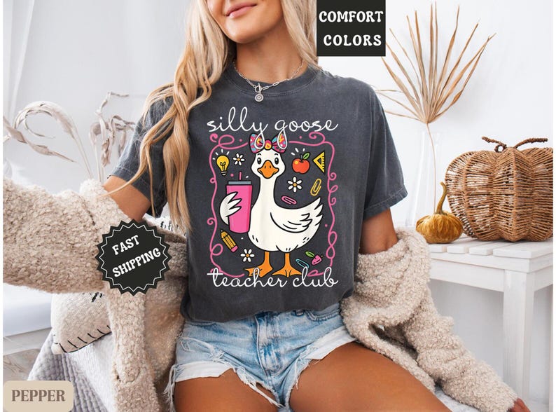 Puede incluir: Camiseta gris oscuro con un gr&aacute;fico caprichoso de un ganso blanco sosteniendo un vaso rosa, rodeado de ilustraciones escolares. El texto dice "silly goose teacher club". La camiseta es de color confort.
