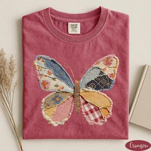 May include: Crimson-colored t-shirt with a crew neck. The shirt features a large butterfly applique made of various patterned fabrics, including floral, denim, and gingham. The butterfly is centered on the front of the shirt.