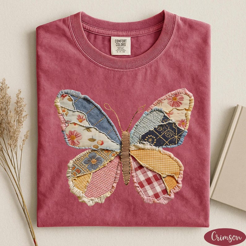 May include: Crimson-colored t-shirt with a crew neck. The shirt features a large butterfly applique made of various patterned fabrics, including floral, denim, and gingham. The butterfly is centered on the front of the shirt.
