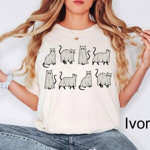 May include: A white t-shirt with a graphic of seven cats dressed as ghosts. The text "Ivory" is in the bottom right corner of the image.