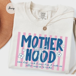 May include: Ivory-colored t-shirt with the word "MOTHERHOOD" in blue, framed by pink stripes. The shirt features a bow graphic and the text "some days I rock it, some days it rocks me either way we're rockin'". A gold necklace rests on the shirt.