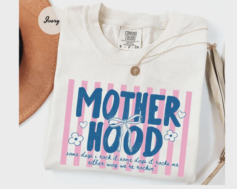 May include: Ivory-colored t-shirt with the word "MOTHERHOOD" in blue, framed by pink stripes. The shirt features a bow graphic and the text "some days I rock it, some days it rocks me either way we're rockin'". A gold necklace rests on the shirt.