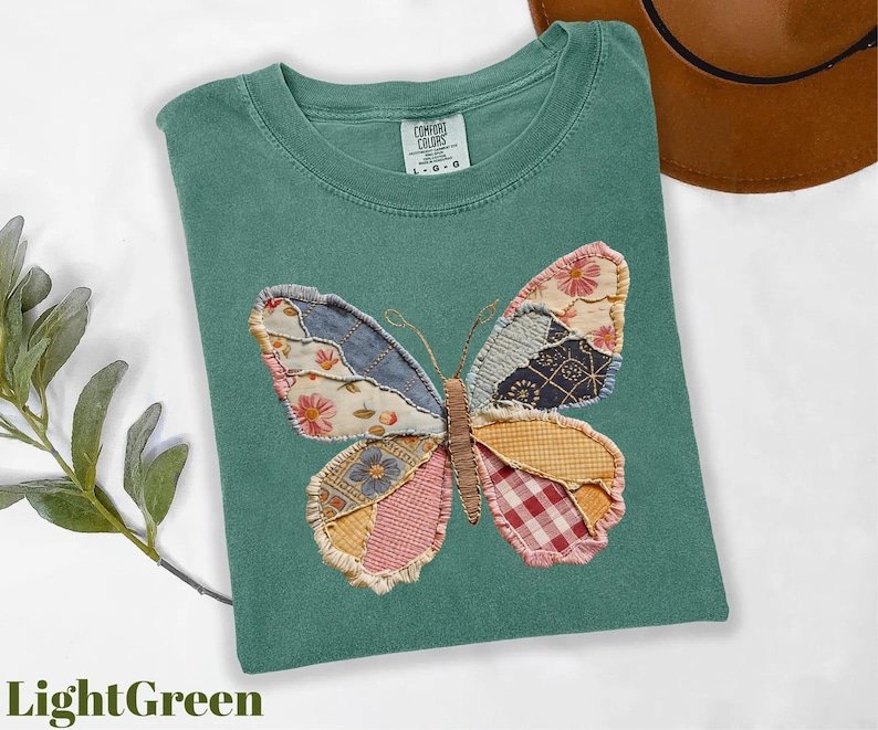 May include: Light green t-shirt with a patchwork butterfly design. The butterfly wings feature various fabric patterns in shades of blue, pink, yellow, and red. The shirt is a Comfort Colors brand, size L-G-O.