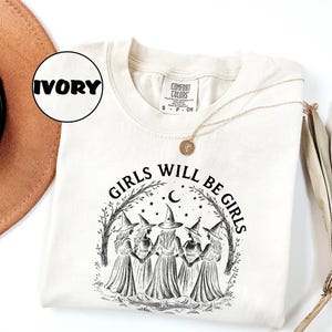 May include: Ivory-coloured sweatshirt with the text "GIRLS WILL BE GIRLS" in black. The graphic features five witch-like figures in dresses and hats, surrounded by a crescent moon, stars, and trees. A gold necklace is draped across the shirt.