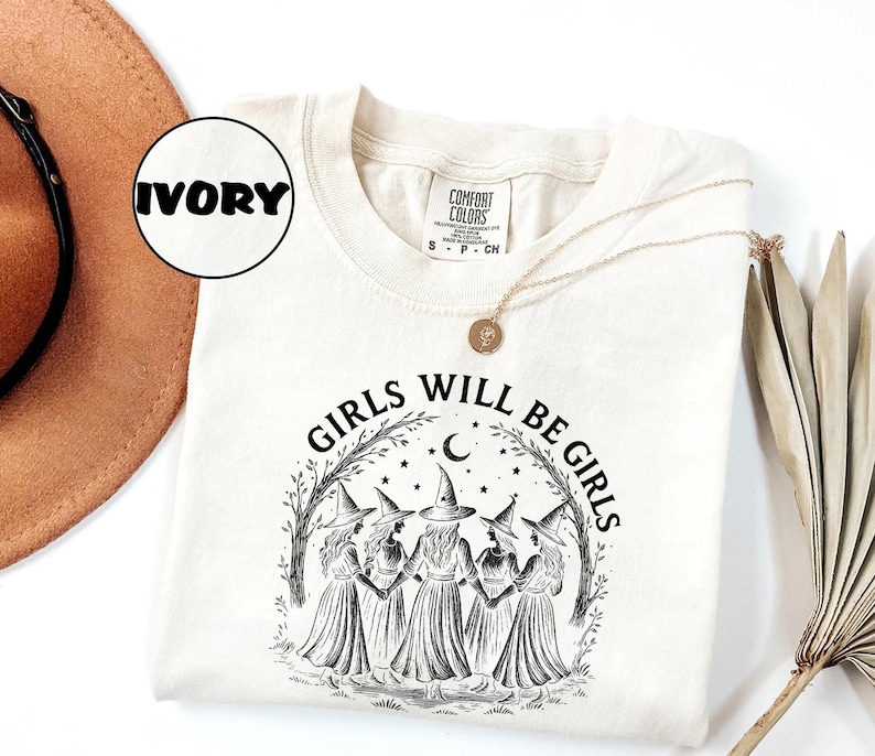 May include: Ivory-coloured sweatshirt with the text "GIRLS WILL BE GIRLS" in black. The graphic features five witch-like figures in dresses and hats, surrounded by a crescent moon, stars, and trees. A gold necklace is draped across the shirt.