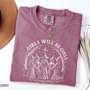 May include: Berry-coloured t-shirt with the text "GIRLS WILL BE GIRLS" in white. The graphic features five figures in witch hats and dresses, holding hands under a crescent moon and stars. A silver necklace with a pendant is on the shirt.