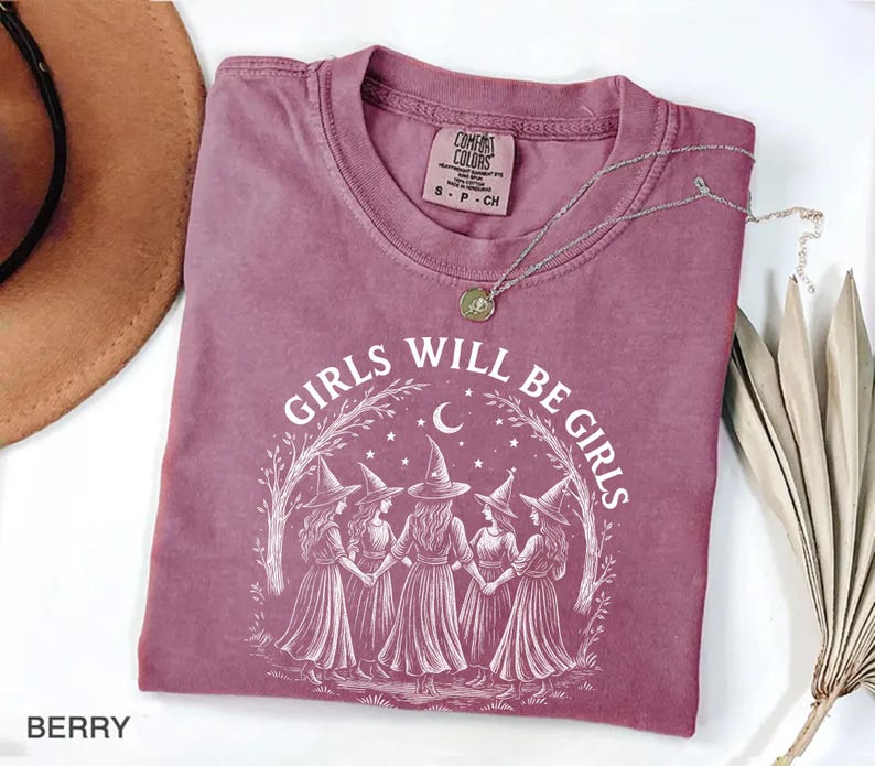May include: Berry-coloured t-shirt with the text "GIRLS WILL BE GIRLS" in white. The graphic features five figures in witch hats and dresses, holding hands under a crescent moon and stars. A silver necklace with a pendant is on the shirt.