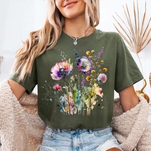 May include: Military green t-shirt featuring a watercolor floral design with purple, pink, and yellow flowers. The graphic includes delicate stems and leaves, creating a soft, artistic look. The shirt is a comfortable, casual style.