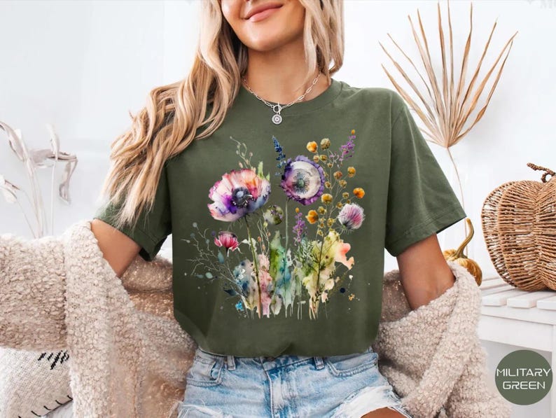 May include: Military green t-shirt featuring a watercolor floral design with purple, pink, and yellow flowers. The graphic includes delicate stems and leaves, creating a soft, artistic look. The shirt is a comfortable, casual style.