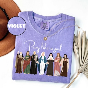 May include: Violet Comfort Colors crewneck sweatshirt with the phrase "Pray like a girl" and illustrations of women. A gold necklace and a brown hat are also in the image. The shirt is a size small.