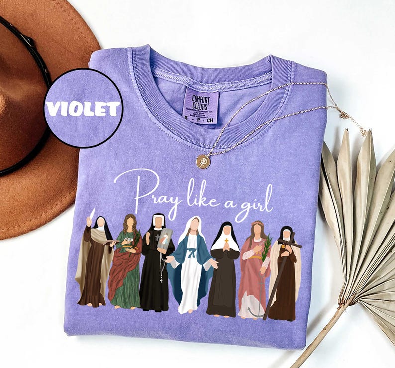 May include: Violet Comfort Colors crewneck sweatshirt with the phrase "Pray like a girl" and illustrations of women. A gold necklace and a brown hat are also in the image. The shirt is a size small.