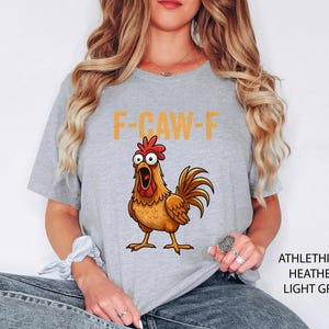 May include: A light grey heather t-shirt featuring a graphic of a brown chicken with wide, surprised eyes and an open beak. Above the chicken, the text F-CAW-F is printed in a bold, yellow, sans-serif font. The shirt has short sleeves and a crew neck.