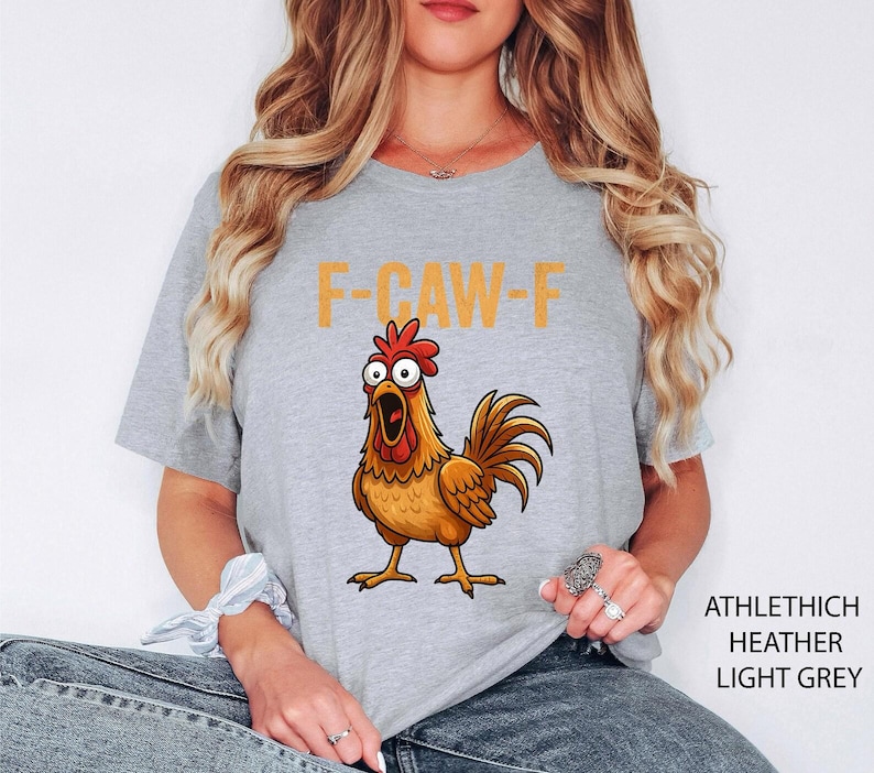 May include: A light grey heather t-shirt featuring a graphic of a brown chicken with wide, surprised eyes and an open beak. Above the chicken, the text F-CAW-F is printed in a bold, yellow, sans-serif font. The shirt has short sleeves and a crew neck.