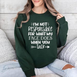 K&ouml;nnte beinhalten: Ein dunkelgr&uuml;ner Pullover mit wei&szlig;em Text, der "Ich bin nicht verantwortlich f&uuml;r das, was mein Gesicht macht, wenn du sprichst" lautet, mit einem wei&szlig;en Pfeil, der nach rechts zeigt.