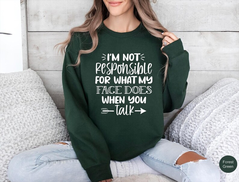 K&ouml;nnte beinhalten: Ein dunkelgr&uuml;ner Pullover mit wei&szlig;em Text, der "Ich bin nicht verantwortlich f&uuml;r das, was mein Gesicht macht, wenn du sprichst" lautet, mit einem wei&szlig;en Pfeil, der nach rechts zeigt.
