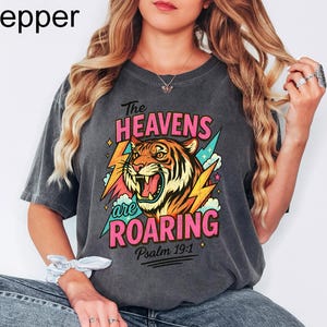 Puede incluir: Camiseta gris oscuro con un gr&aacute;fico de un tigre rugiente, rayos y el texto "The Heavens are Roaring" y "Psalm 19:1". El tigre es naranja, amarillo y negro. El texto es rosa y turquesa.