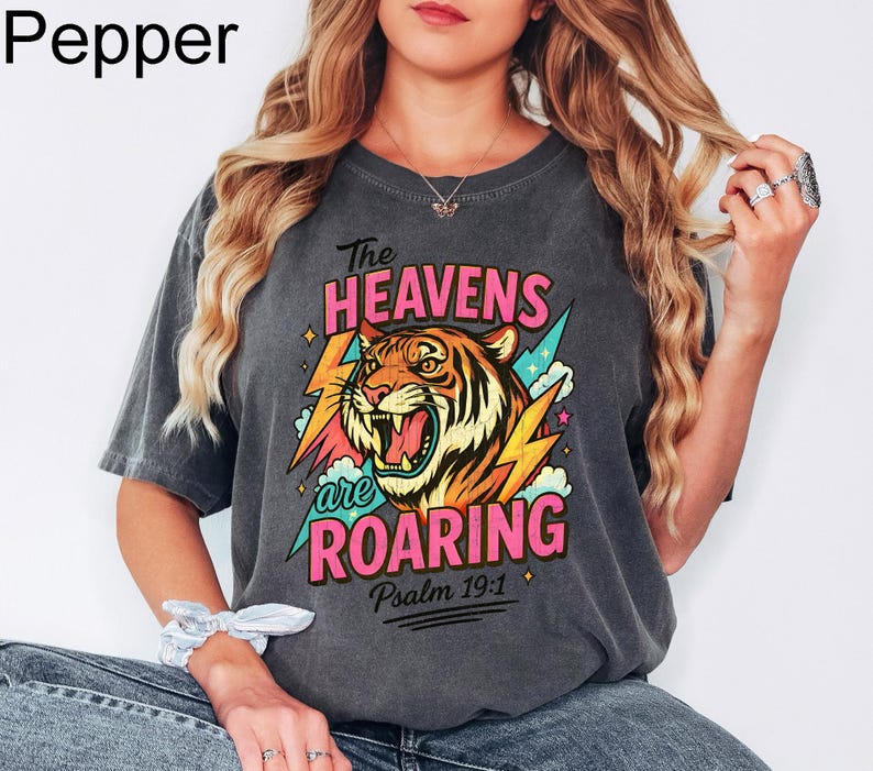 Puede incluir: Camiseta gris oscuro con un gr&aacute;fico de un tigre rugiente, rayos y el texto "The Heavens are Roaring" y "Psalm 19:1". El tigre es naranja, amarillo y negro. El texto es rosa y turquesa.