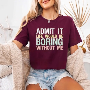 May include: A maroon t-shirt with the text "ADMIT IT LIFE WOULD BE BORING WITHOUT ME" in white and teal lettering. The shirt is short-sleeved and appears to be made of a soft material. The model is wearing denim shorts and a cream-colored cardigan.