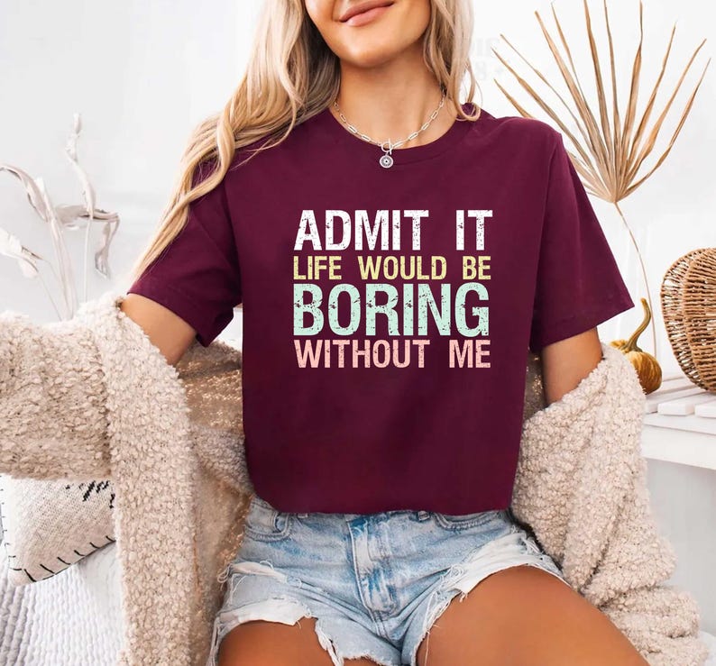 May include: A maroon t-shirt with the text "ADMIT IT LIFE WOULD BE BORING WITHOUT ME" in white and teal lettering. The shirt is short-sleeved and appears to be made of a soft material. The model is wearing denim shorts and a cream-colored cardigan.