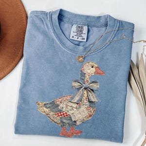 Peut inclure: T-shirt bleu clair Comfort Colors avec un motif d'oie en patchwork et un n&oelig;ud &agrave; carreaux. L'oie est faite de divers motifs de tissu, dont des motifs floraux et en jean. Un collier en or avec un pendentif repose sur le t-shirt.