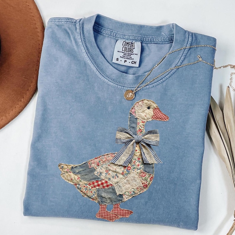 Peut inclure: T-shirt bleu clair Comfort Colors avec un motif d'oie en patchwork et un n&oelig;ud &agrave; carreaux. L'oie est faite de divers motifs de tissu, dont des motifs floraux et en jean. Un collier en or avec un pendentif repose sur le t-shirt.