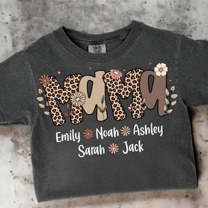 May include: Dark gray t-shirt with the word "MAMA" in a leopard print design, adorned with floral accents. Below, names like Emily, Noah, and Ashley are displayed. A Comfort Colors tag is visible.