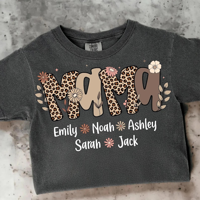 May include: Dark gray t-shirt with the word "MAMA" in a leopard print design, adorned with floral accents. Below, names like Emily, Noah, and Ashley are displayed. A Comfort Colors tag is visible.