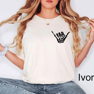 Pode incluir: Uma camiseta branca com uma m&atilde;o de esqueleto preta fazendo um sinal shaka. O texto "Marfim" &eacute; vis&iacute;vel no canto inferior direito da imagem.