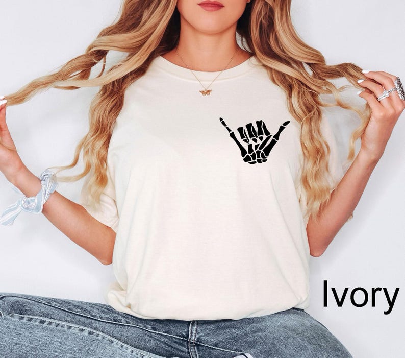 Pode incluir: Uma camiseta branca com uma m&atilde;o de esqueleto preta fazendo um sinal shaka. O texto "Marfim" &eacute; vis&iacute;vel no canto inferior direito da imagem.