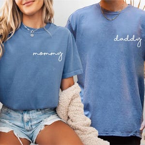 Pu&ograve; includere: Due magliette blu a maniche corte con girocollo in tessuto slavato color denim. La maglietta a sinistra presenta la parola mommy in corsivo bianco sul petto, mentre la maglietta a destra presenta la parola daddy in corsivo bianco.