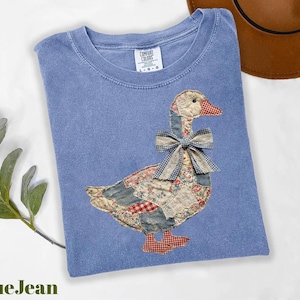 Peut inclure: T-shirt bleu jean avec un motif d'oie en patchwork. L'oie est orn&eacute;e d'un n&oelig;ud et est faite de divers tissus &agrave; motifs. Le t-shirt a un col rond et des manches courtes. Le texte "BlueJean" est en bas &agrave; gauche.