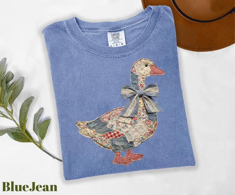Peut inclure: T-shirt bleu jean avec un motif d'oie en patchwork. L'oie est orn&eacute;e d'un n&oelig;ud et est faite de divers tissus &agrave; motifs. Le t-shirt a un col rond et des manches courtes. Le texte "BlueJean" est en bas &agrave; gauche.