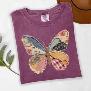 May include: A purple Comfort Colors t-shirt featuring a patchwork butterfly design. The butterfly wings are made of various patterned fabrics in shades of blue, pink, yellow, and red. The shirt is folded and has a tag in the upper center.