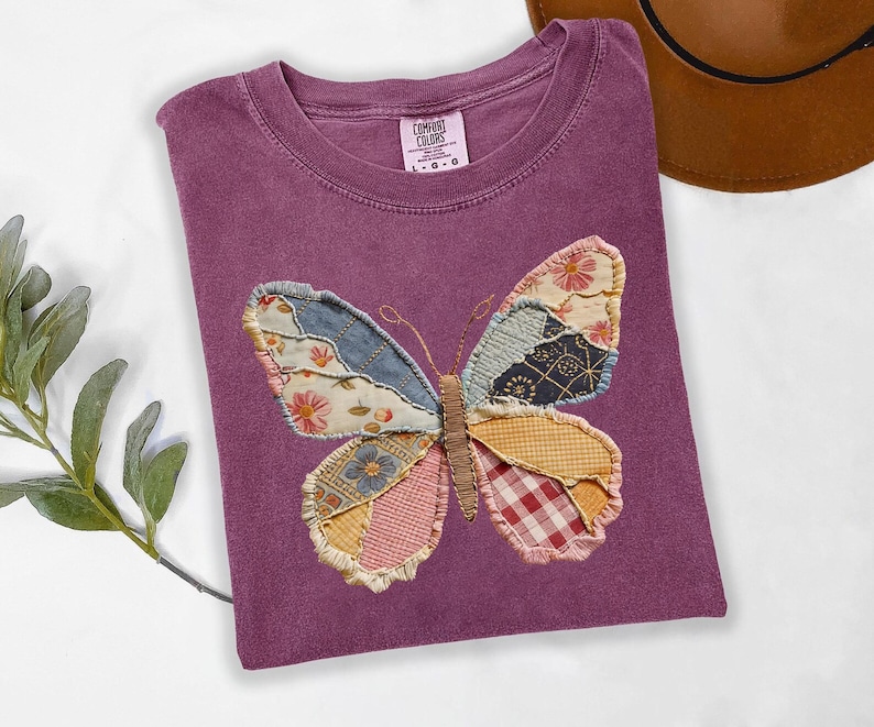 May include: A purple Comfort Colors t-shirt featuring a patchwork butterfly design. The butterfly wings are made of various patterned fabrics in shades of blue, pink, yellow, and red. The shirt is folded and has a tag in the upper center.