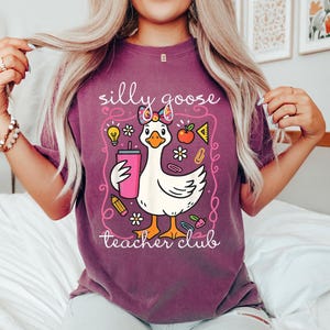Puede incluir: Camiseta granate con un dise&ntilde;o de ganso de dibujos animados sosteniendo una bebida rosa. El ganso est&aacute; enmarcado con las palabras "silly goose teacher club". El dise&ntilde;o incluye gr&aacute;ficos escolares como una manzana, un l&aacute;piz y una bombilla.