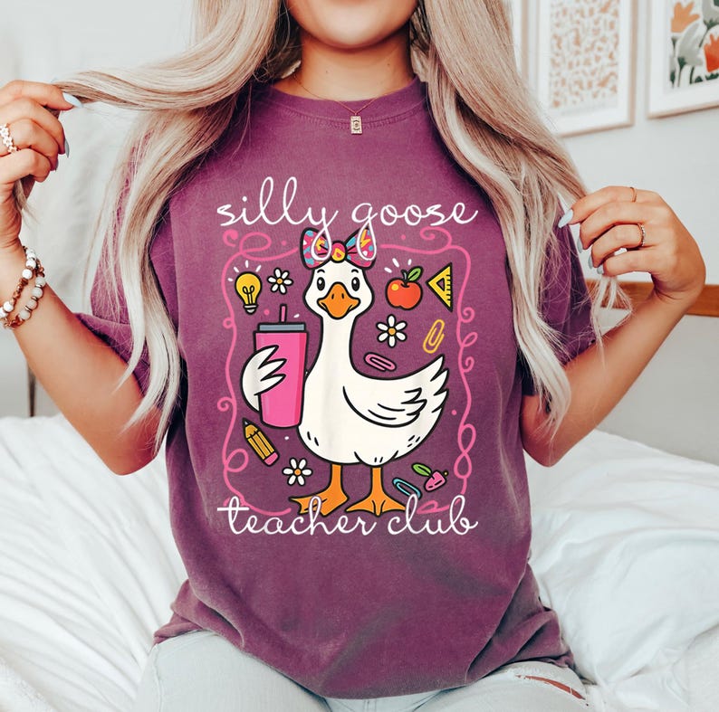 Puede incluir: Camiseta granate con un dise&ntilde;o de ganso de dibujos animados sosteniendo una bebida rosa. El ganso est&aacute; enmarcado con las palabras "silly goose teacher club". El dise&ntilde;o incluye gr&aacute;ficos escolares como una manzana, un l&aacute;piz y una bombilla.