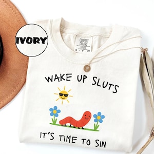 Puede incluir: Camiseta Comfort Colors color marfil con el texto "WAKE UP SLUTS IT'S TIME TO SIN". El dise&ntilde;o incluye un sol sonriente, un gusano rojo y flores azules. Un collar dorado est&aacute; sobre la camiseta.