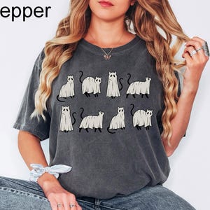 May include: A dark gray t-shirt with a graphic print of nine white ghost cats. The cats are in various poses, some are standing, some are sitting, and some are walking. The shirt has a relaxed fit and a crew neck.