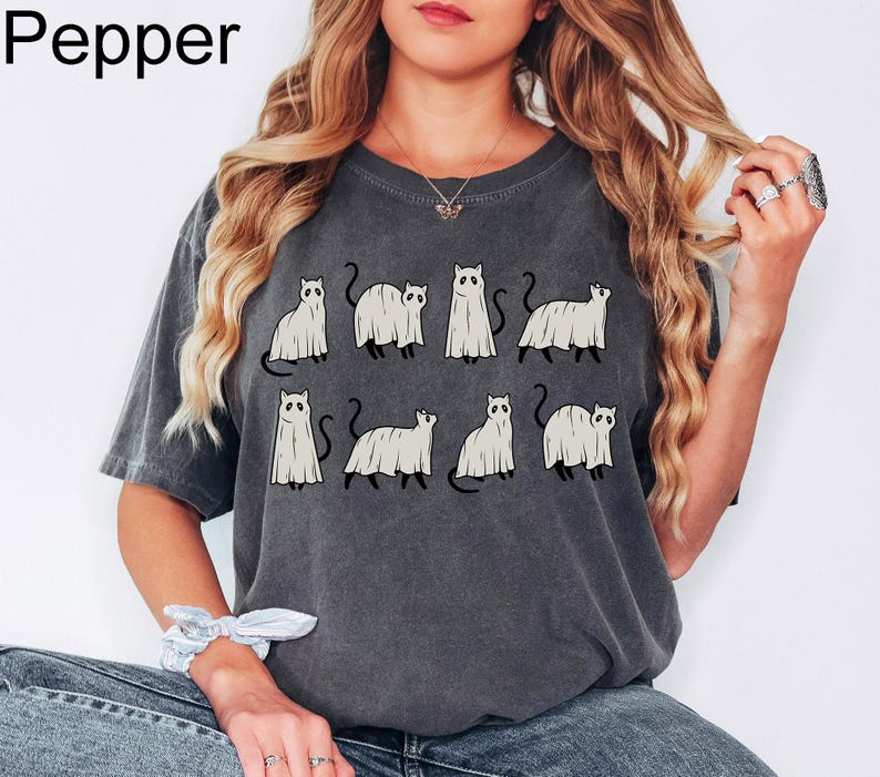May include: A dark gray t-shirt with a graphic print of nine white ghost cats. The cats are in various poses, some are standing, some are sitting, and some are walking. The shirt has a relaxed fit and a crew neck.