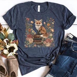 K&ouml;nnte beinhalten: Ein dunkelblau meliertes Kurzarm-T-Shirt mit einer zentralen Grafik eines Rotfuchses, der auf einem Stapel alter B&uuml;cher sitzt, umgeben von einem farbenfrohen Arrangement aus verschiedenen Blumen und Bl&auml;ttern in Orange-, Rosa- und Wei&szlig;t&ouml;nen.