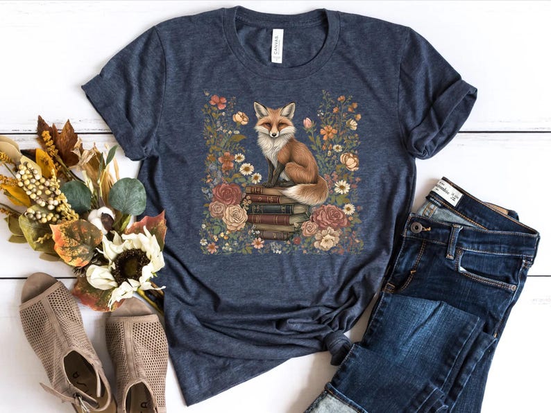 K&ouml;nnte beinhalten: Ein dunkelblau meliertes Kurzarm-T-Shirt mit einer zentralen Grafik eines Rotfuchses, der auf einem Stapel alter B&uuml;cher sitzt, umgeben von einem farbenfrohen Arrangement aus verschiedenen Blumen und Bl&auml;ttern in Orange-, Rosa- und Wei&szlig;t&ouml;nen.