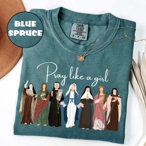 May include: Blue Spruce Comfort Colors t-shirt with the phrase "Pray like a girl" and an illustration of seven women. A gold necklace with a pendant is on the shirt. The shirt is a size S-P-CH.