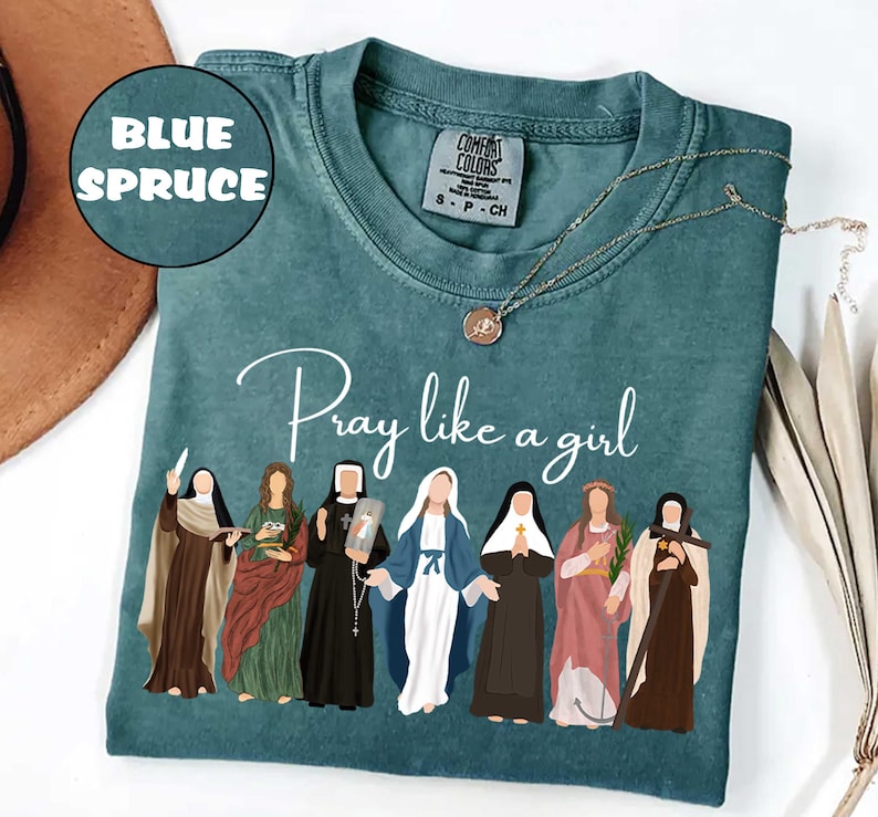 May include: Blue Spruce Comfort Colors t-shirt with the phrase "Pray like a girl" and an illustration of seven women. A gold necklace with a pendant is on the shirt. The shirt is a size S-P-CH.